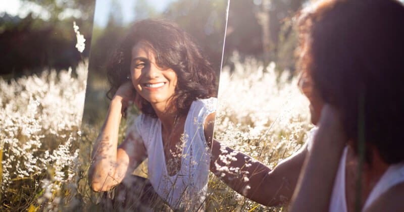 A woman with dark hair sits in a sunlit field, smiling at her reflection in a mirror among tall grass and wildflowers, embodying the simple joys of how to be happy.