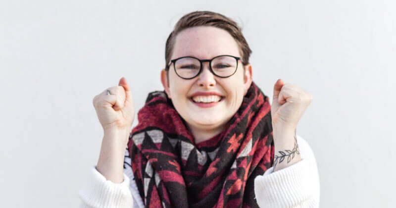 Person with short hair, glasses, and a patterned scarf smiles and raises both fists in the air against a plain white background, perfectly capturing how to be happy in a simple everyday moment.
